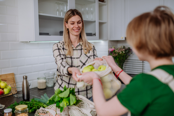 A mother unpacking local food in zero waste packaging from bag with help of daughter in kitchen at home.