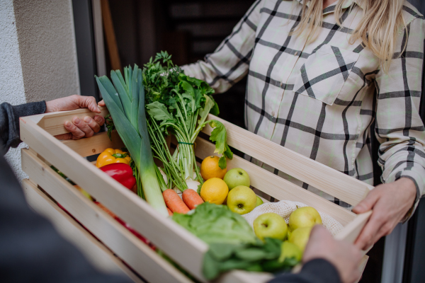 A man holding crate with vegetales and fruit and delivering it to woman standing at doorway.