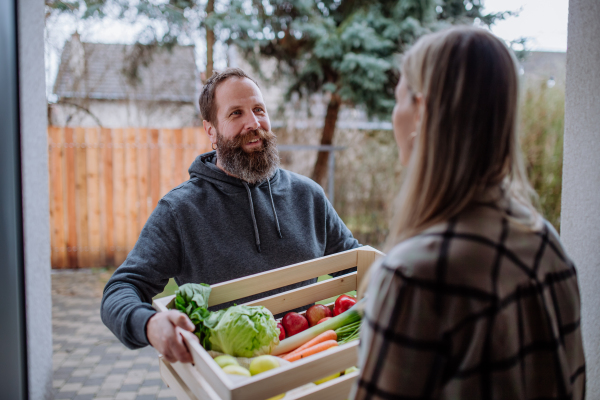 A mature man holding crate with vegetales and fruit and delivering it to woman standing at doorway.