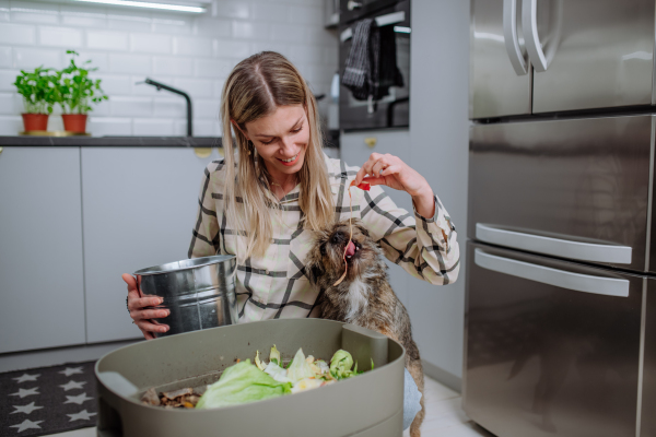 A woman throwing vegetable cuttings in a compost bucket in kitchen and feeding dog.