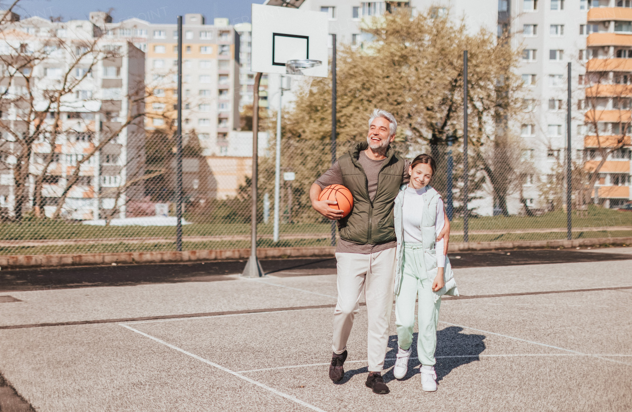 A happy father and teen daughter embracing and looking at camera outside at basketball court.