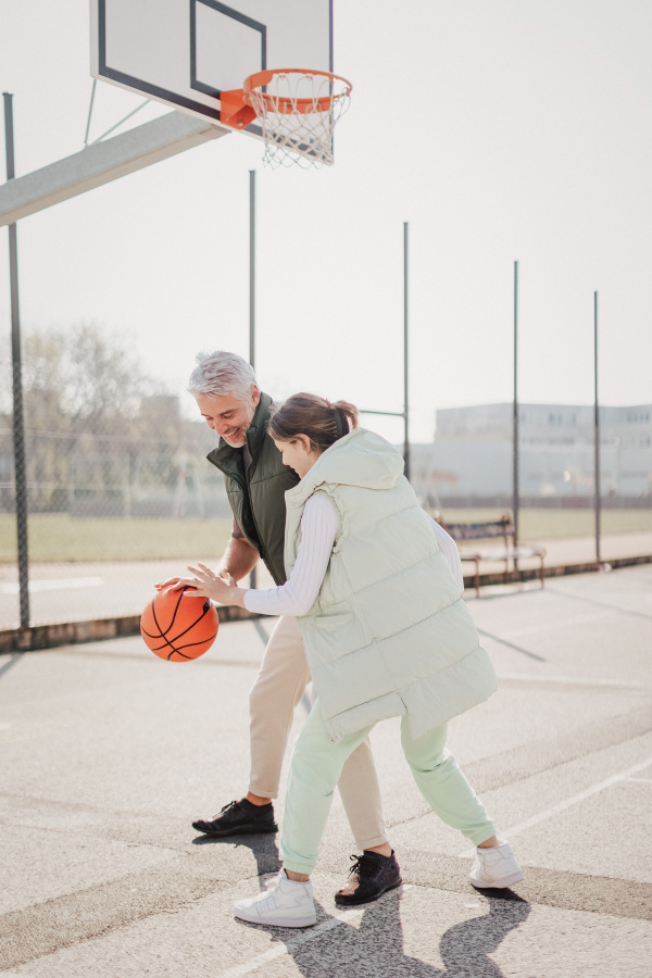 A happy father and teen daughter playing basketball outside at court.