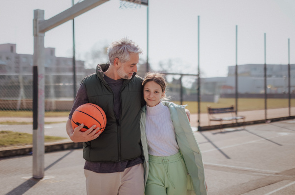 A happy father and teen daughter embracing and looking at camera outside at basketball court.