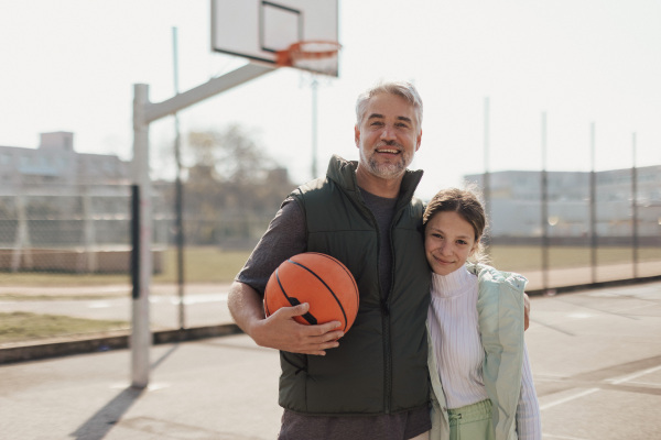 A happy father and teen daughter embracing and looking at camera outside at basketball court.