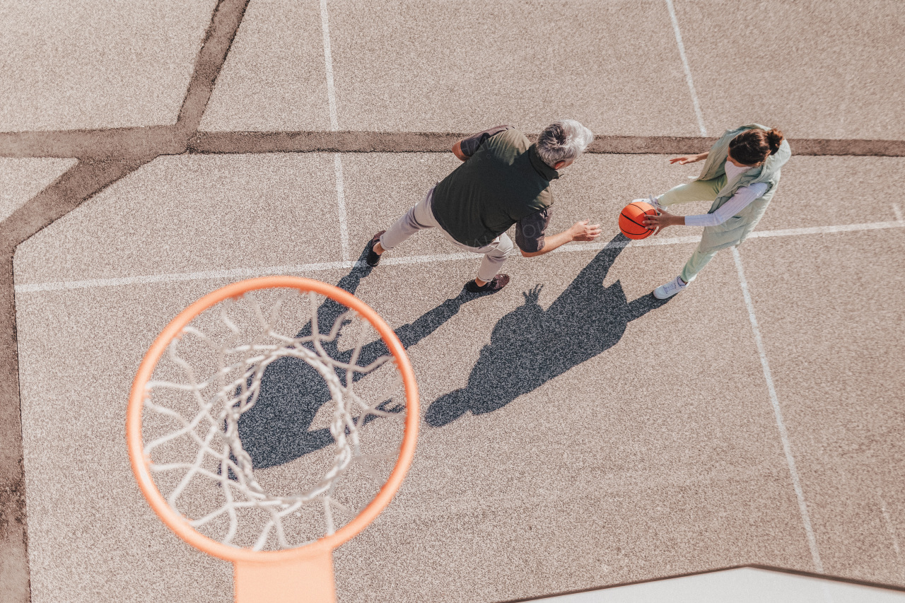 A father and teenage daughter playing basketball outside at court, high angle view above hoop net.