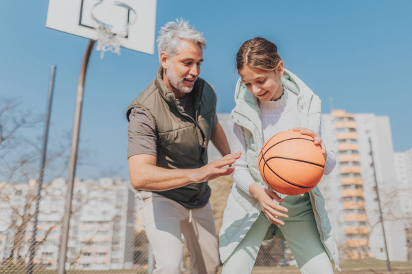 A happy father and teen daughter playing basketball outside at court.