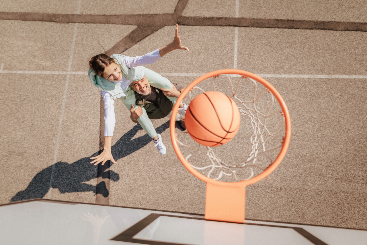 A father and teenage daughter playing basketball outside at court, high angle view above hoop net.