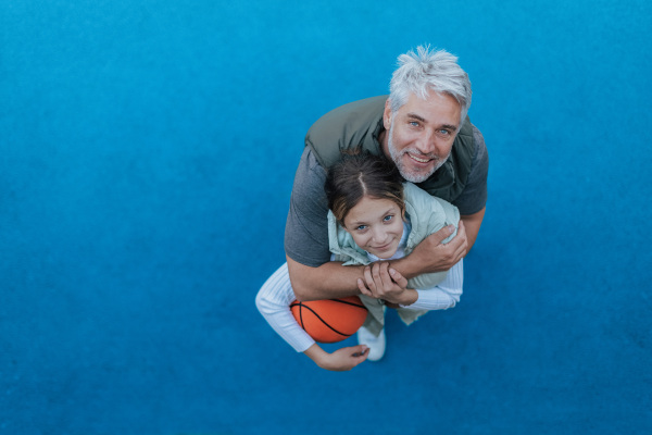 A father and teenage daughter playing basketball outside at court, high angle view.