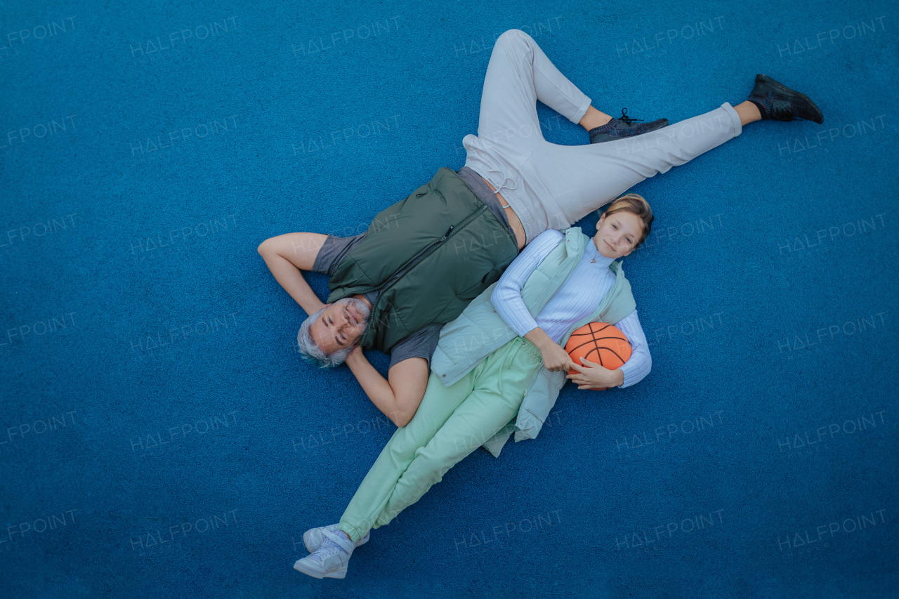 A happy father and teen daughter sitting, embracing and looking at camera outside at basketball court.