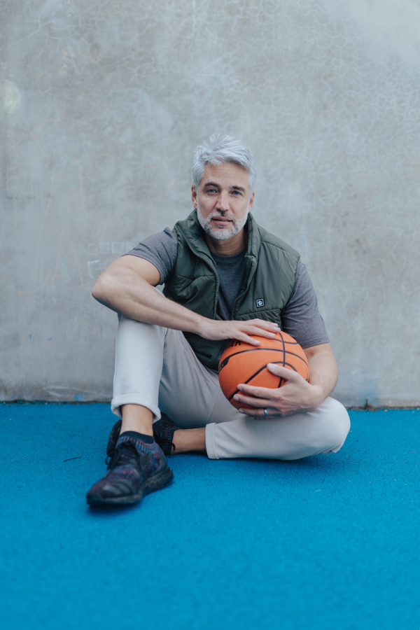 A mature man with baskatball sitting on courtoutdoors and looking at camera.