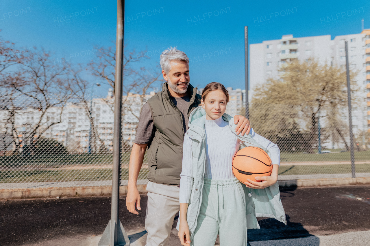 A happy father and teen daughter embracing and looking at camera outside at basketball court.