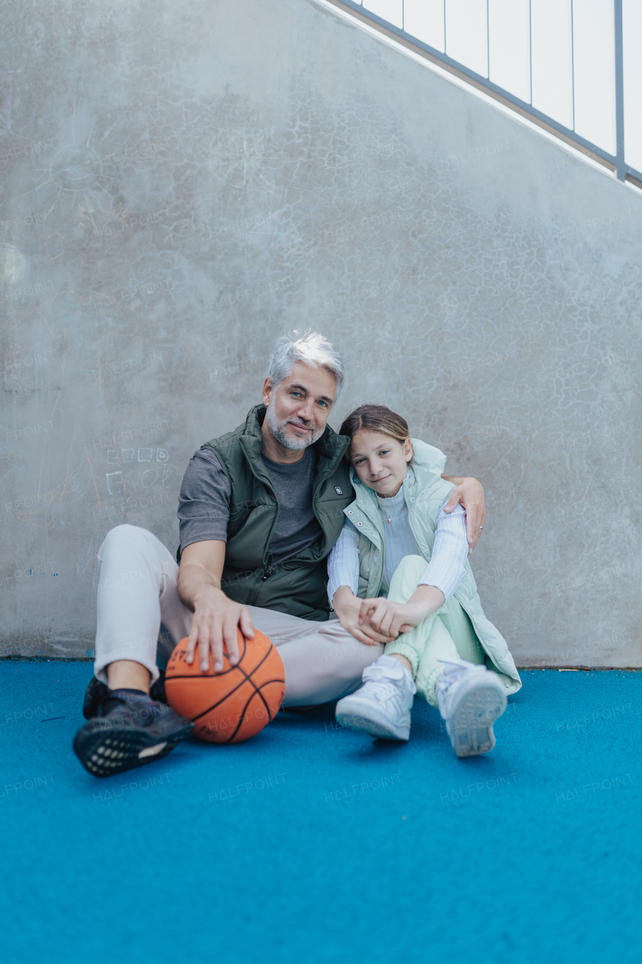 A happy father and teen daughter sitting, embracing and looking at camera outside at basketball court.