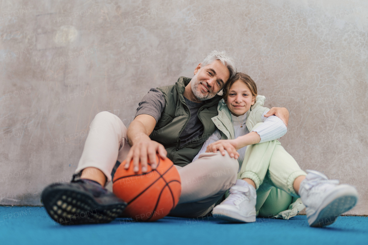 A happy father and teen daughter sitting, embracing and looking at camera outside at basketball court.