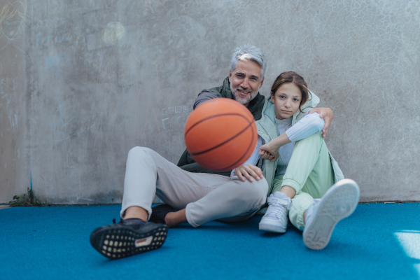 A happy father and teen daughter embracing and looking at camera outside at basketball court.