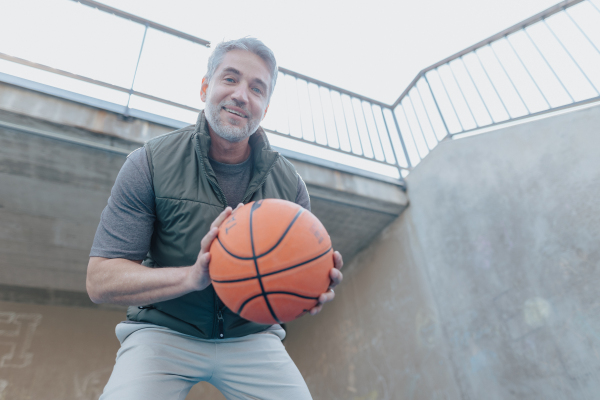 A mature man with baskatball sitting on courtoutdoors and looking at camera.