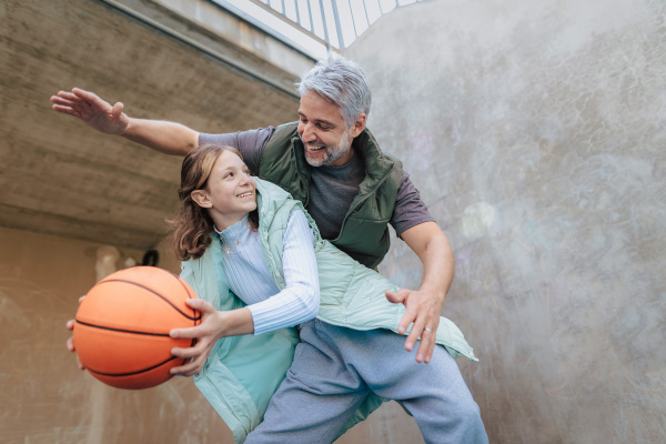 A happy father and teen daughter playing basketball outside at court.