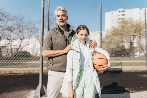 A happy father and teen daughter embracing and looking at camera outside at basketball court.