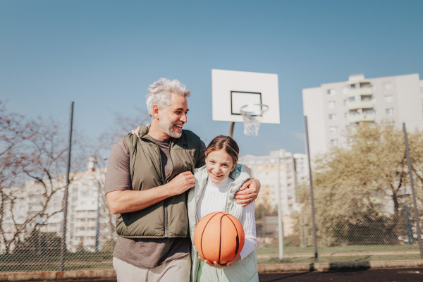 A happy father and teen daughter embracing and looking at camera outside at basketball court.