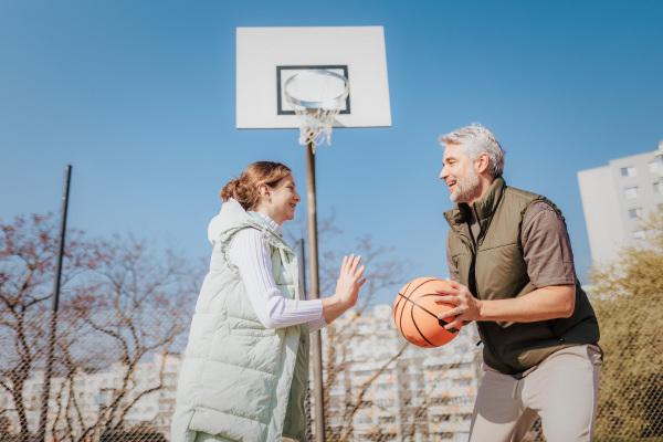 A happy father and teen daughter playing basketball outside at court.