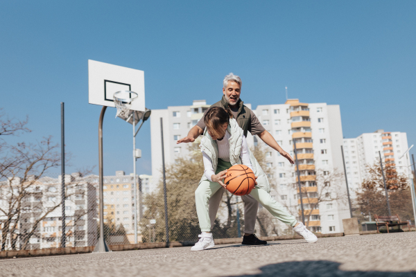 A happy father and teen daughter embracing and looking at camera outside at basketball court.