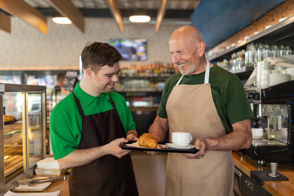 A happy waiter with Down syndrome serving coffee with help of his collegue at cafe.