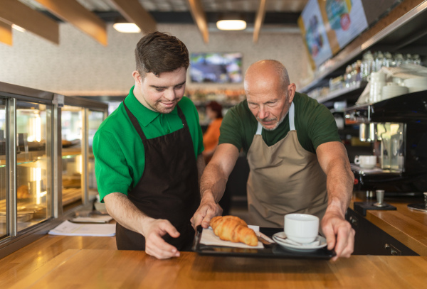 A happy waiter with Down syndrome serving coffee with help of his collegue at cafe.