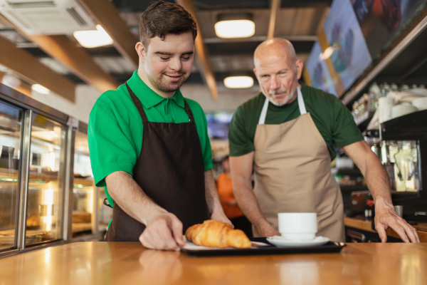 A happy waiter with Down syndrome serving coffee with help of his collegue at cafe.