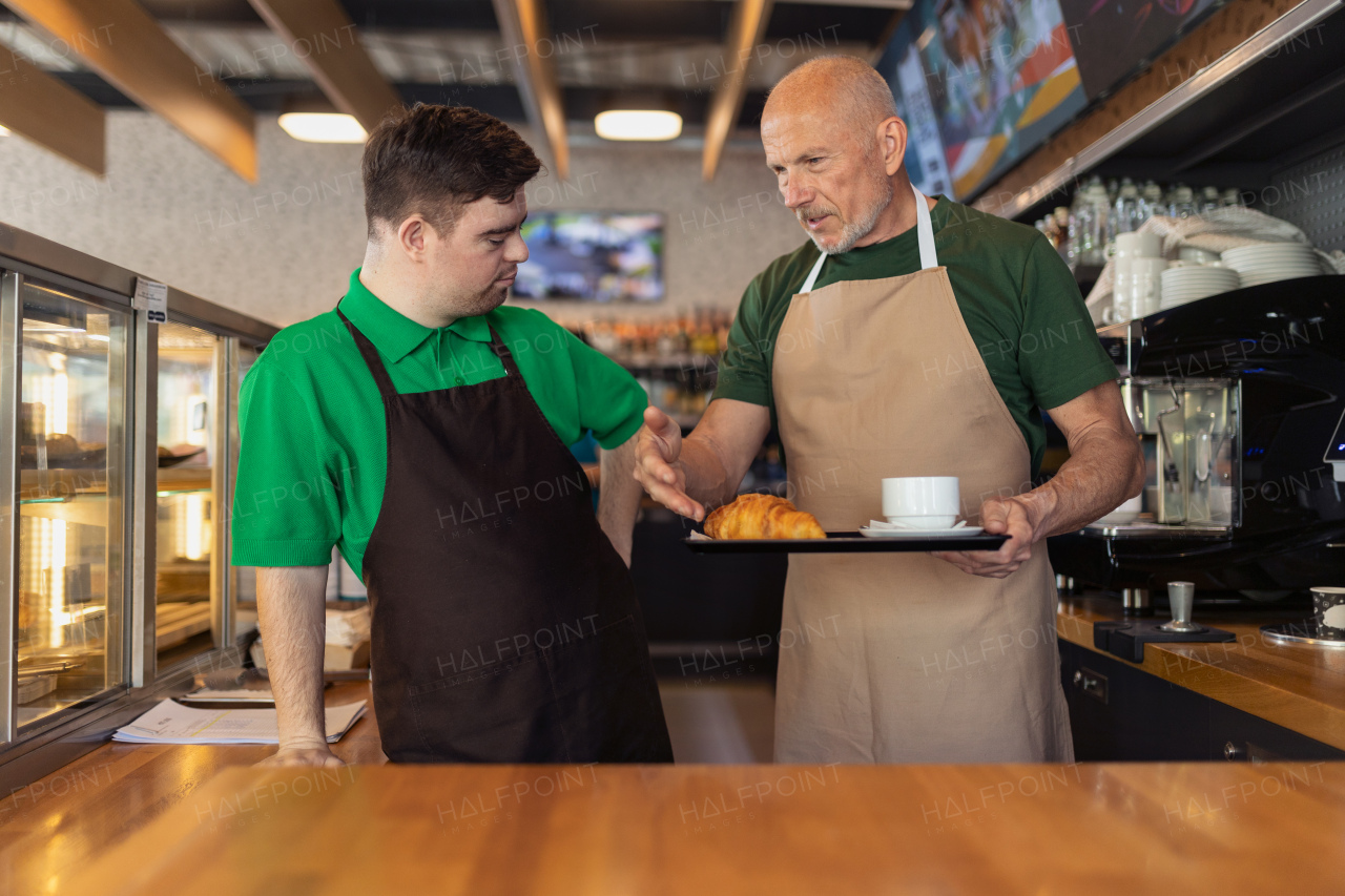 A happy waiter with Down syndrome serving coffee with help of his collegue at cafe.