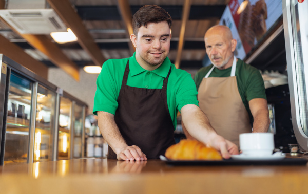 A happy waiter with Down syndrome serving coffee with help of his collegue at cafe.