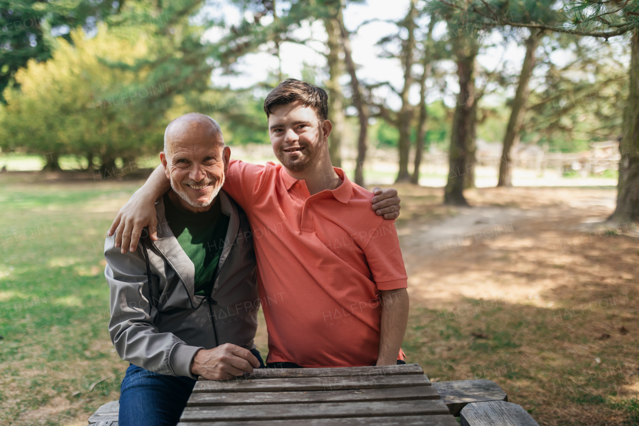 A happy senior father with his young son with Down syndrome embracing and sitting in park.