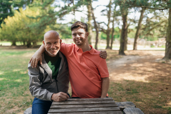 A happy senior father with his young son with Down syndrome embracing and sitting in park.