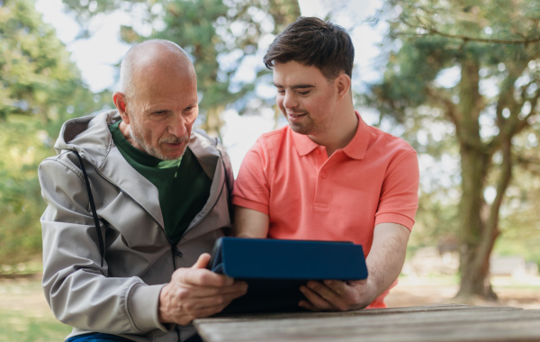 A happy senior father with his young son with Down syndrome using tablet and sitting in park.