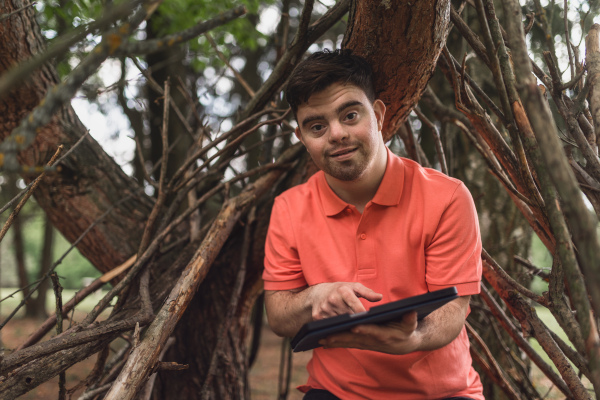 A portrait of happy young man with down syndrome standing outdoors in park, using digital tablet.