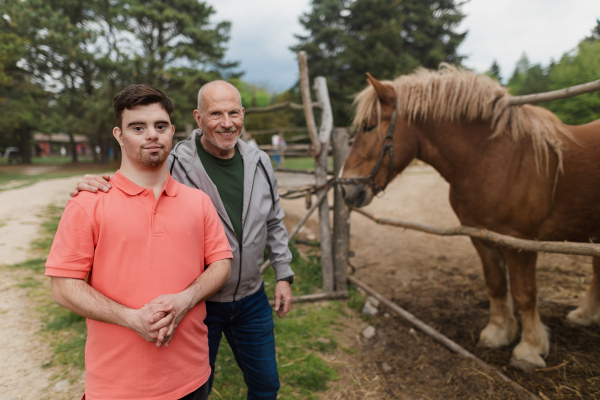 A happy senior father with his adult son with Down syndrome at ranch looking at horse.