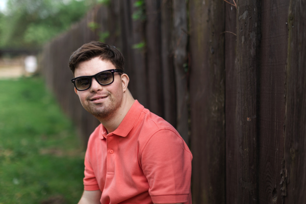 A portrait of happy young man with down syndrome standing outdoors in park and wearing sunglasses