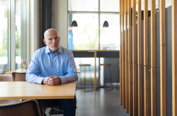 A sad senior man sitting at empty table with blurred cafe interior in background