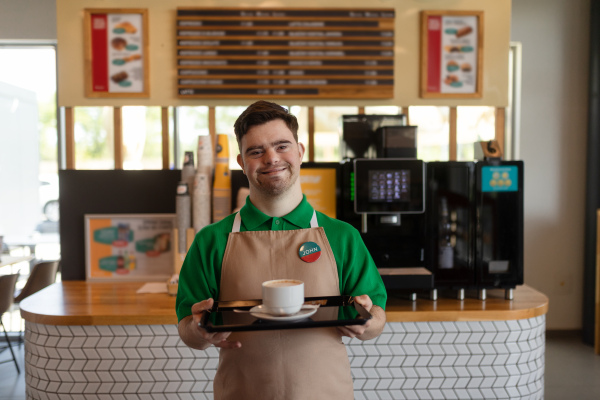 A happy waiter with Down syndrome carrying coffee to customer in cafe at gas station. Social inclusion concept.