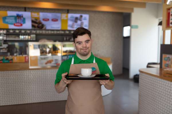 A happy waiter with Down syndrome carrying coffee to customer in cafe at gas station. Social inclusion concept.