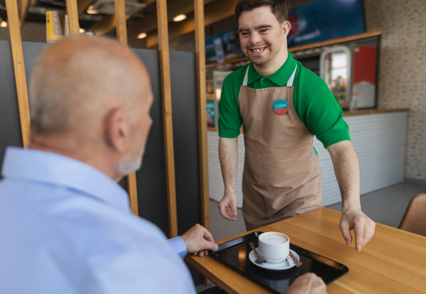 A happy waiter with Down syndrome serving coffee to senior customer at cafe.