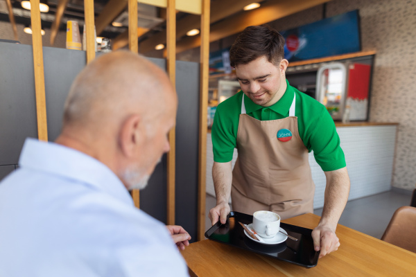 A happy waiter with Down syndrome serving coffee to senior customer at cafe.