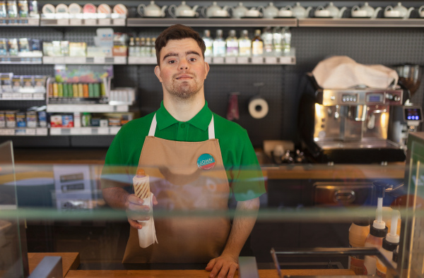 A happy waiter with Down syndrome serving baguette and passing it to costumer in cafe at gas station.