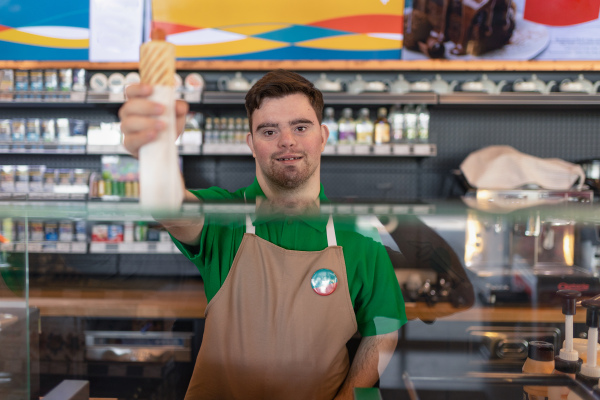 A happy waiter with Down syndrome serving baguette and passing it to costumer in cafe at gas station.