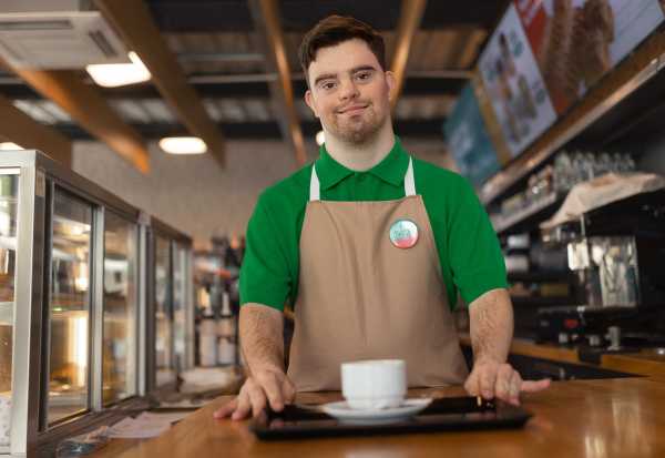 A happy waiter with Down syndrome carrying coffee to customer in cafe at gas station. Social inclusion concept.