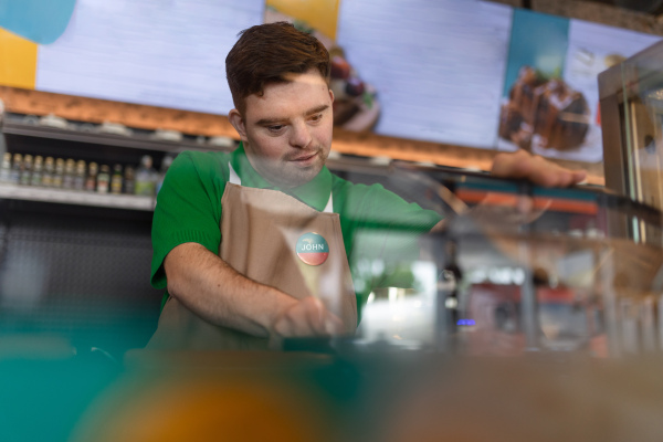 A happy waiter with Down syndrome prepairing hot-dog to customer in cafe at gas station.