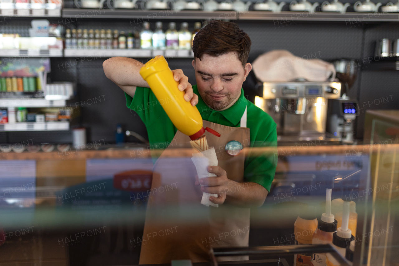 A happy waiter with Down syndrome prepairing hot-dog to customer in cafe at gas station.