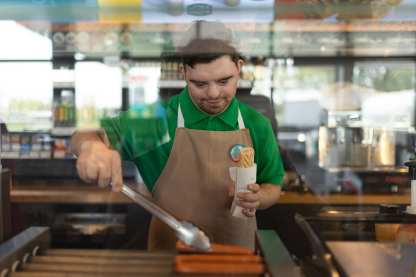 A happy waiter with Down syndrome prepairing hot-dog to customer in cafe at gas station.