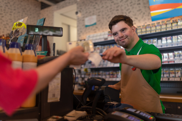 A cheerful young Down Syndrome employee taking cash payment from costumer in gas station cafe.