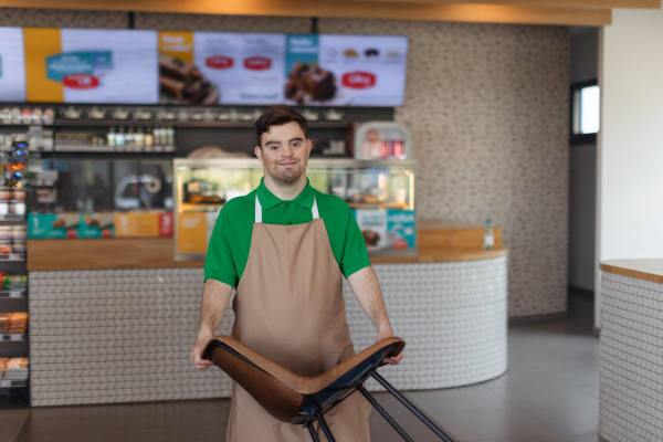 A happy waiter with Down syndrome carrying chair in cafe at gas station. Social inclusion concept.