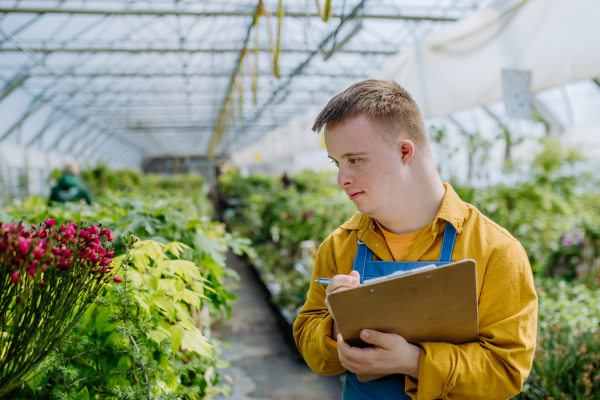 A young man with Down syndrome working in garden centre, holidng clipboard and checking plants.