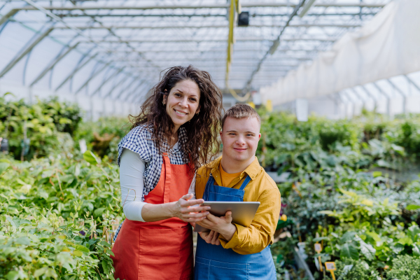 An experienced woman florist helping young employee with Down syndrome to check flowers on tablet in garden centre.